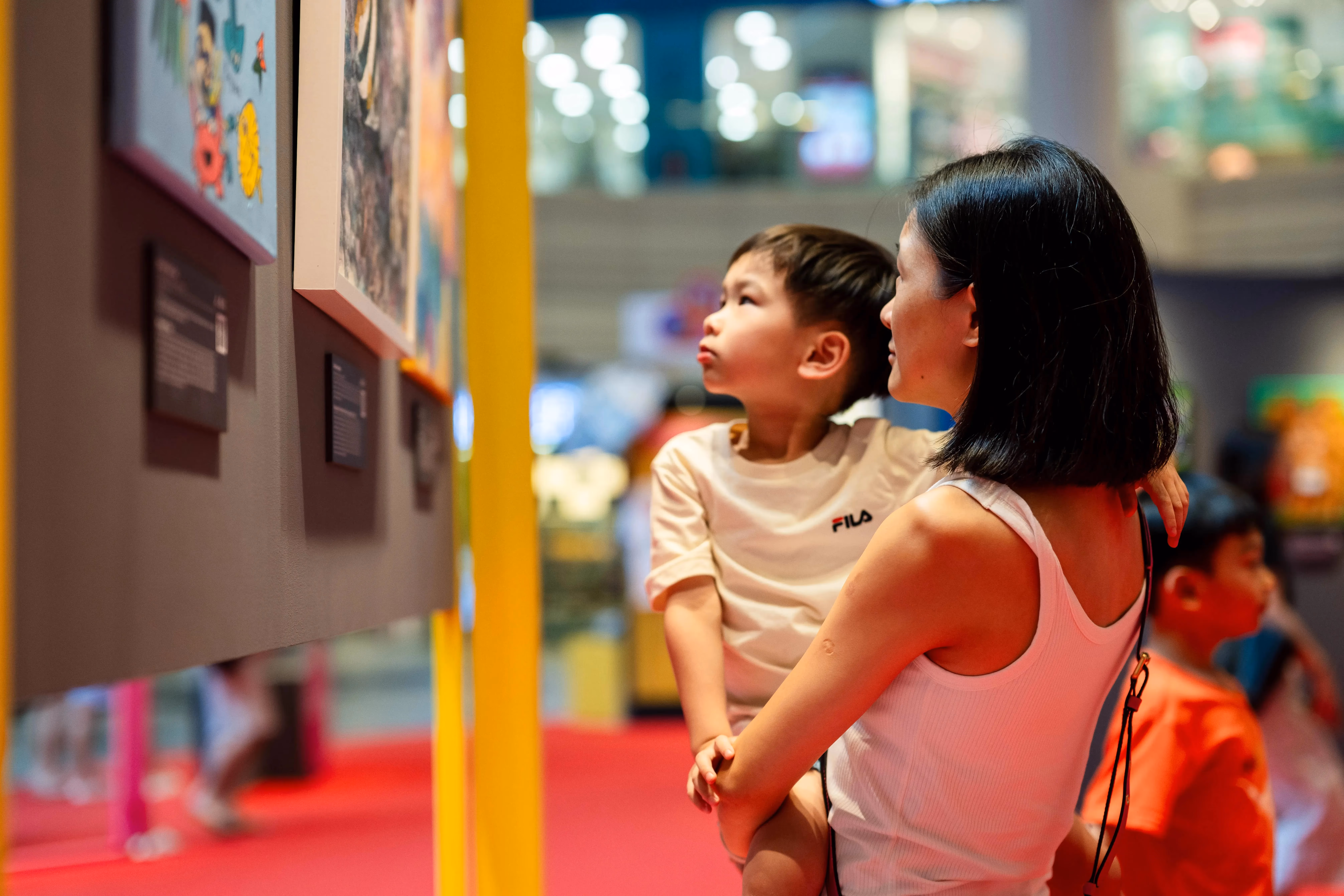 A woman holding a child is observing colorful artwork in a brightly lit gallery with red flooring.